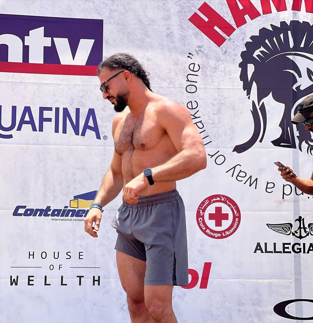 Shirtless man in athletic shorts standing in front of a backdrop with various sponsor logos.